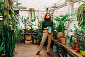 Caring red haired female holding a jug between plants growing in pots placed on shelf in indoor garden with various houseplants