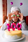Optimistische ältere Frau mit Spielzeugaugen, die sich amüsiert, während sie an einem Tisch mit einem Kuchen sitzt, der mit Kerzen mit der Zahl 100 verziert ist und ein Jubiläum feiert