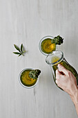 Top view of crop anonymous persons hand pouring fresh marijuana infusion into glass with herb placed on white table