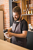Adult bearded male barista in apron wiping moka pot with napkin while standing at counter in cafe