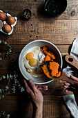 From above unrecognizable male cook using wooden spoon to mix pumpkin puree with eggs and flour in bowl while preparing pie on timber table near cutting board and towel