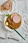 Top view of spoon placed in bowl of yummy lentil soup served with slices of sausage and leaf of parsley on marble table and gray napkin