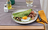 Homemade breakfast of spinach pancakes with bacon, egg and mushrooms served on a white plate with a salt and oil shaker on a checkered tablecloth.