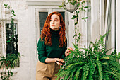 Serious female with ginger hair trimming green branch of potted fern growing in indoor garden