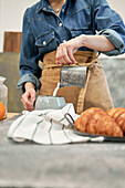 Young female blogger pouring milk from jug into cup against photo camera at table with croissants at home