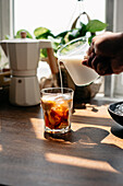Crop unrecognizable person pouring milk into glass cup with freshly brewed coffee and ice cube on wooden table in light kitchen