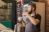 Side view of bearded male barista in apron standing at bar table with cup of hot drink and giving order to client in coffee shop