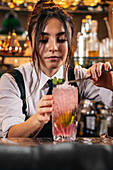 Young woman in uniform putting mint leaves on top of strawberry mojito while working in bar at night