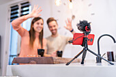 Positive couple of bloggers waving hand to cellphone with microphone while sitting at kitchen counter with cup of coffee at home