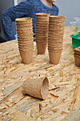 Cropped unrecognizable child sitting at table with pile of cardboard cups and containers against gardening shovel and fork while looking away