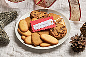 Teller mit verschiedenen süßen Keksen und Geschenkanhänger auf einem Tisch mit Weihnachtsdekoration von oben