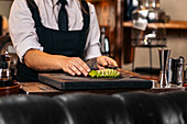 Crop woman in uniform chopping fresh lime on counter while preparing cocktail during work in bar