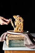 Anonymous person eating cut panettone located with kitchen towel on plate placed on stack of withered books against dark backdrop