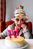 Fröhliche ältere Frau mit Sonnenbrille und lustigem Hut beißt in eine Kerze, während sie ihr 100-jähriges Jubiläum mit einem Geburtstagskuchen feiert und in die Kamera schaut