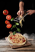 Cook grating cheese while tomatoes and basil leaves falling on raw pizza against black background