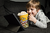 Little boy lying down comfortable on sofa with popcorn bucket while eating and watching cartoon on tablet