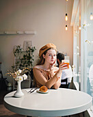 French female in beret sitting at table in cafe with aromatic glass of coffee and freshly baked croissant