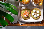 From above of delicious avocado toasts with salmon and burrata cheese served on table with glasses of fresh juice and herbal tea placed near exotic Monstera deliciosa plant