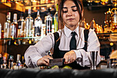 Young woman in uniform chopping fresh lime on counter while preparing cocktail during work in bar