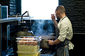 Young man in grey apron tasting food from a big metal pan on restaurant kitchen