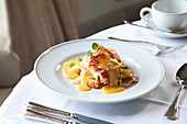 Appetitliche Eier Benedict mit Speck und Sauce Hollandaise, serviert in einem weißen Teller auf einem Tisch mit einer Tasse Kaffee und Besteck in einem Restaurant während des Brunchs