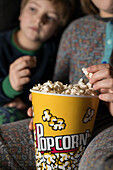 Cropped blurred young amazed siblings kids sitting comfortable on sofa with popcorn bucket while eating and watching TV
