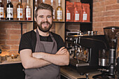 Bärtiger positiver männlicher Barista in Schürze, der die Arme verschränkt, während er in der Nähe von Geräten und Regalen mit Glasflaschen in einem Kaffeehaus steht und in die Kamera schaut