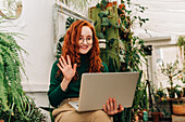 Positiv eingestellte rothaarige Mitarbeiterin mit Brille sitzt zwischen Zimmerpflanzen und winkt der Netbook-Kamera während einer Videokonferenz zu