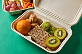 From above of lunch boxes with healthy food including crackers carrot sticks grapes cherry tomatoes with kiwi broccoli walnut and tangerine on green background