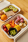 From above of lunch boxes with healthy food including crackers carrot sticks grapes cherry tomatoes with kiwi broccoli walnut and tangerine on yellow background