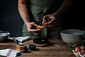 Unrecognizable guy in apron breaking raw egg over bowl while preparing pastry on lumber table near kitchenware