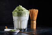 Glass of fresh dalgona coffee with matcha and Japanese tea ceremony accessories consisting of bamboo whisk called chasen and traditional scoop named chashaku placed on gray surface