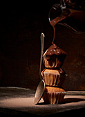 Sweet chocolate topping pouring on tasty naked muffins served on top of each other near spoon on black background in room