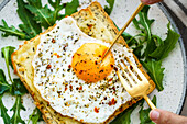 From above cropped unrecognizable person eating toast with eggs and cheese and rocket lettuce served on plate on table background