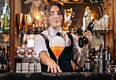 Young woman in uniform using smoke gun to create bubble on top of tequila sunrise cocktail during work in bar