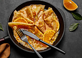 Von oben hausgemachte Crêpes Suzette mit Orangenlikör und Orangenscheiben auf einem Teller mit Messer und Gabel auf einer Betonfläche serviert