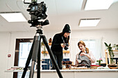 Woman taking photo of chocolate muffins on digital camera against blogger talking during cooking process in kitchen