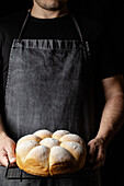 Crop unrecognizable baker standing at counter with fresh bread buns placed on baking pan in kitchen