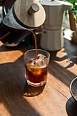 Crop unrecognizable person pouring freshly brewed coffee from glass coffee press into glass with ice cube on wooden table in light kitchen