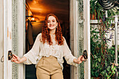 Charming young female with long ginger hair opening door of indoor garden with various potted houseplants on shelves