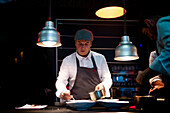 Cook holding spoon and little pan while adding sauce in plate on restaurant kitchen