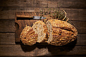 Top view of appetizing freshly baked homemade natural bread loaf with crispy crust and oat flakes on wooden table with knife and aromatic herbs
