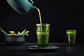 Gesunder japanischer Matcha-Tee, der während einer Teezeremonie aus einer grünen Teekanne in ein Glas gegossen wird, vor schwarzem Hintergrund