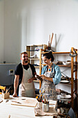 Positive middle aged female and young male in casual clothes and aprons smiling while creating clay plate during pottery class in workshop