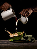Crop unrecognizable person pouring splashing milk and freshly brewed coffee into green cup on table in kitchen on black background