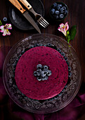 Delicious blueberry mousse cake with purple cream decorated with fresh berries served on glass stand on dark table with flowers