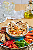 From above of bowl with hummus and tortilla chips surrounded with various vegetables and croutons served on wicker mat near olive oil and lemonade on timber table