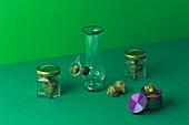 Komposition einer runden Glasbong neben einer trockenen Cannabispflanze in einer Schale auf grünem Untergrund
