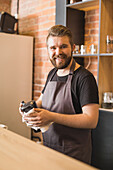 Adult bearded male barista in apron wiping moka pot with napkin while standing at counter in cafe