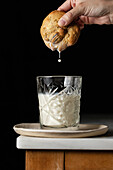 Crop anonymous person dipping fresh sweet cookie into glass of milk placed on corner of table against black background in kitchen